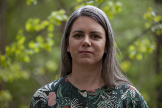 Porträtt på Signe Tonér utomhus i blomstermönstrad tunika mot abstrakt grönskande bakgrund.