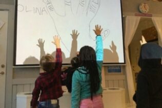 Barn sträcker upp händerna mot en projicerad bild av en hand med namntext, som del av ett matematikutforskande i förskolan.