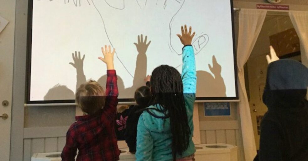 Barn sträcker upp händerna mot en projicerad bild av en hand med namntext, som del av ett matematikutforskande i förskolan.
